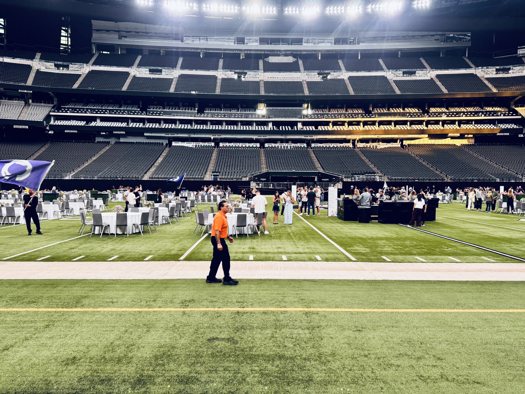Dining tables laid out across the turf at Allegiant Stadium, with empty tiered seating rising into the dark above.