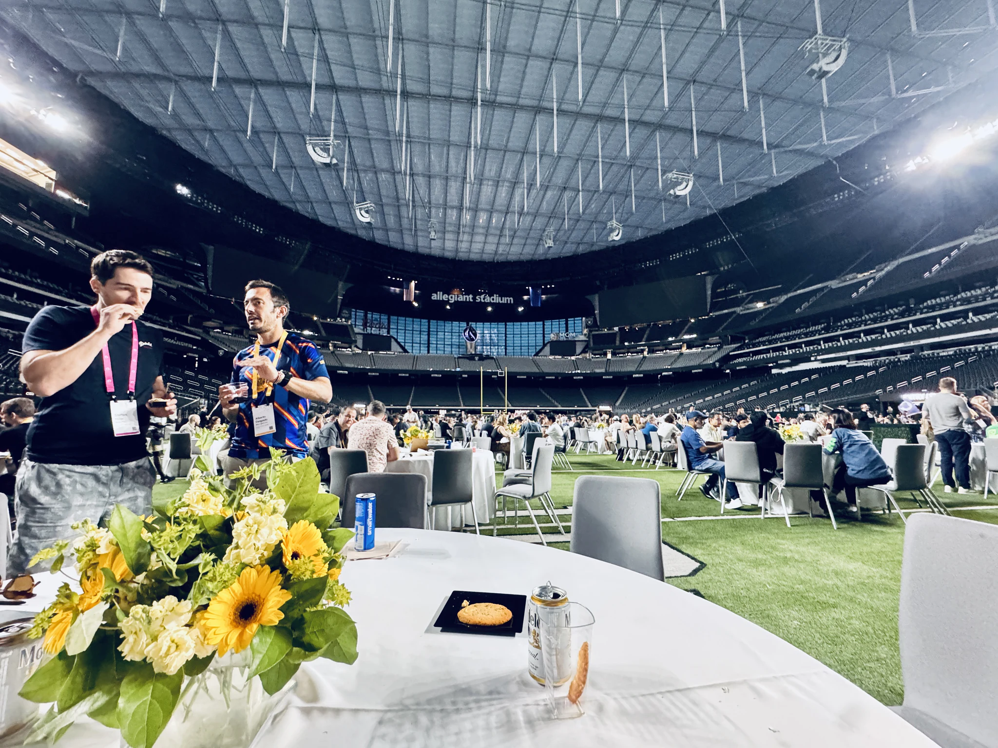 Two people holding drinks beside a table of sunflowers, the Allegiant Stadium scoreboard and lattice roof behind them.