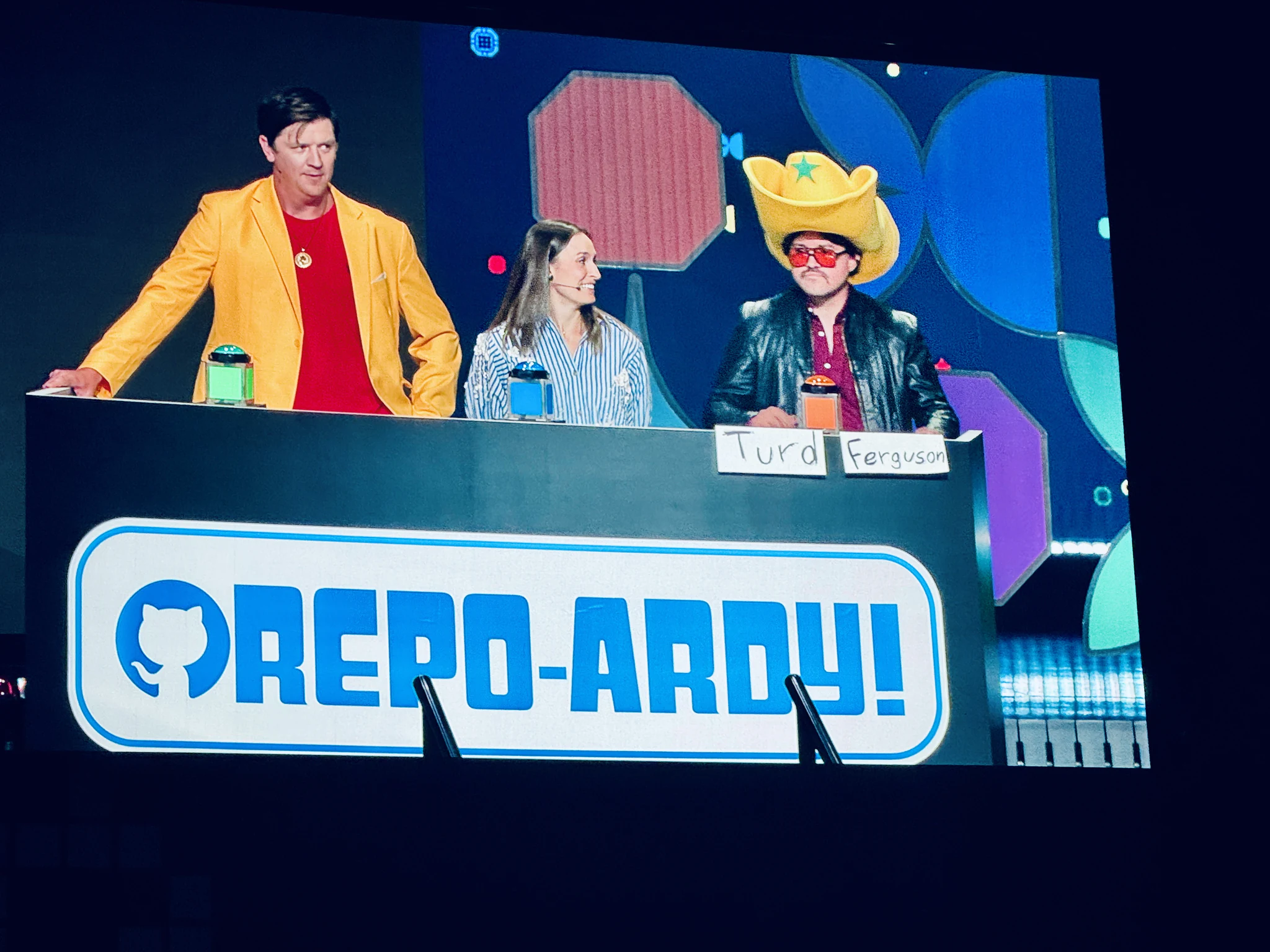 Three contestants behind a game show podium on a huge screen, beneath a GitHub-branded Repo-ardy! sign.