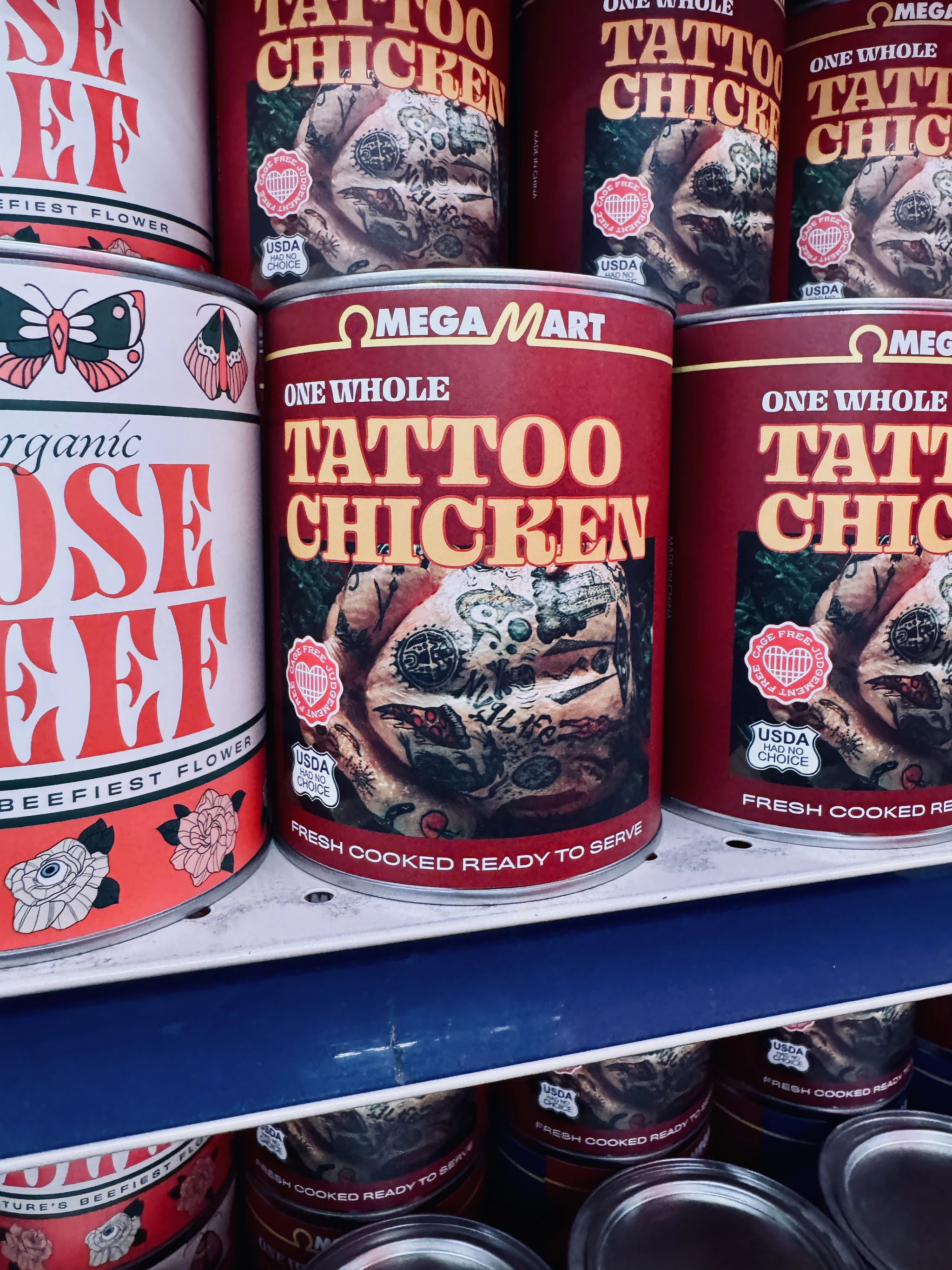 Shelves of tins in Omega Mart labelled One Whole Tattoo Chicken and Organic Rose Beef.