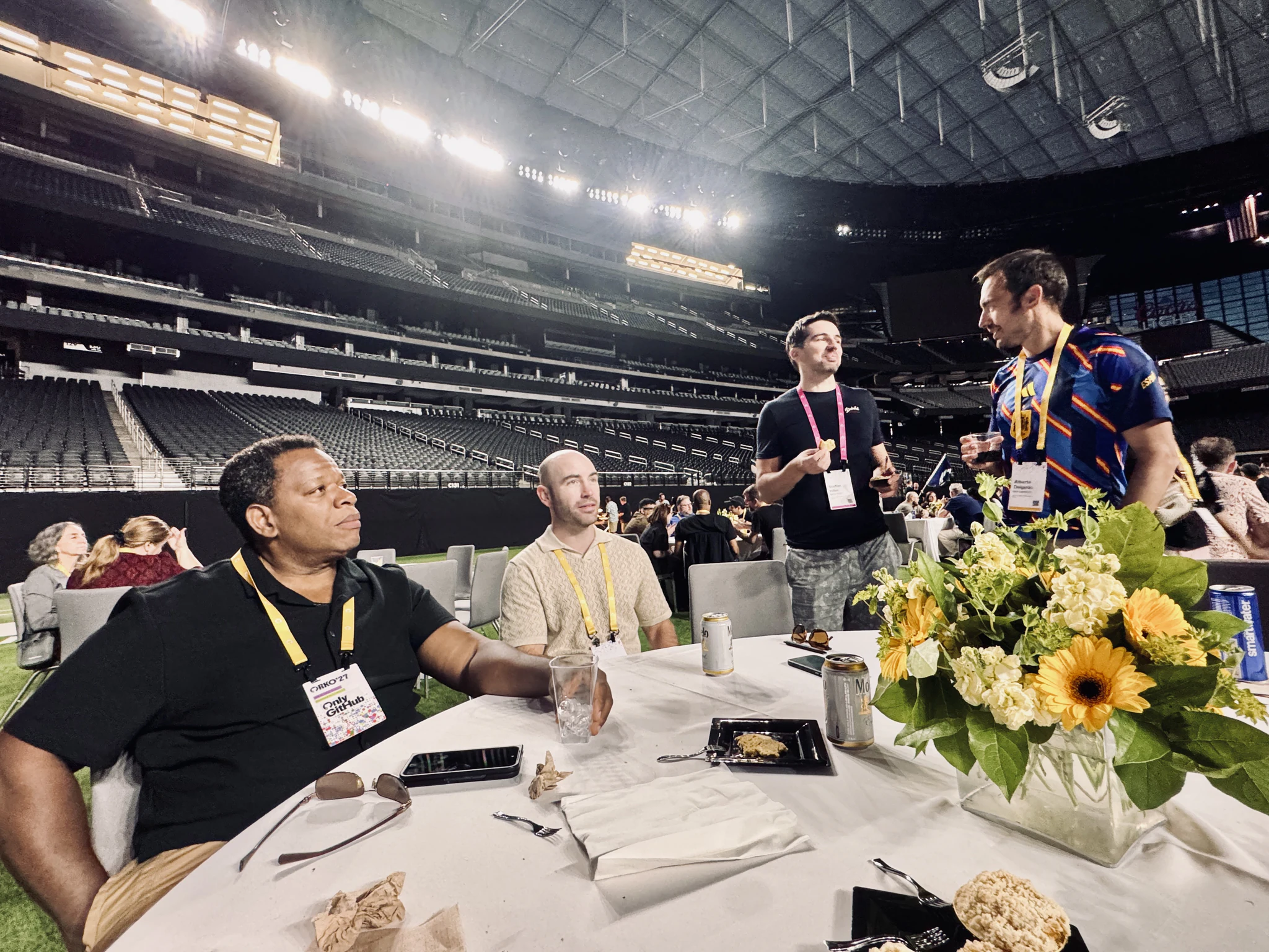 Colleagues sitting and standing around a round table on the stadium turf, a vase of sunflowers in the foreground.