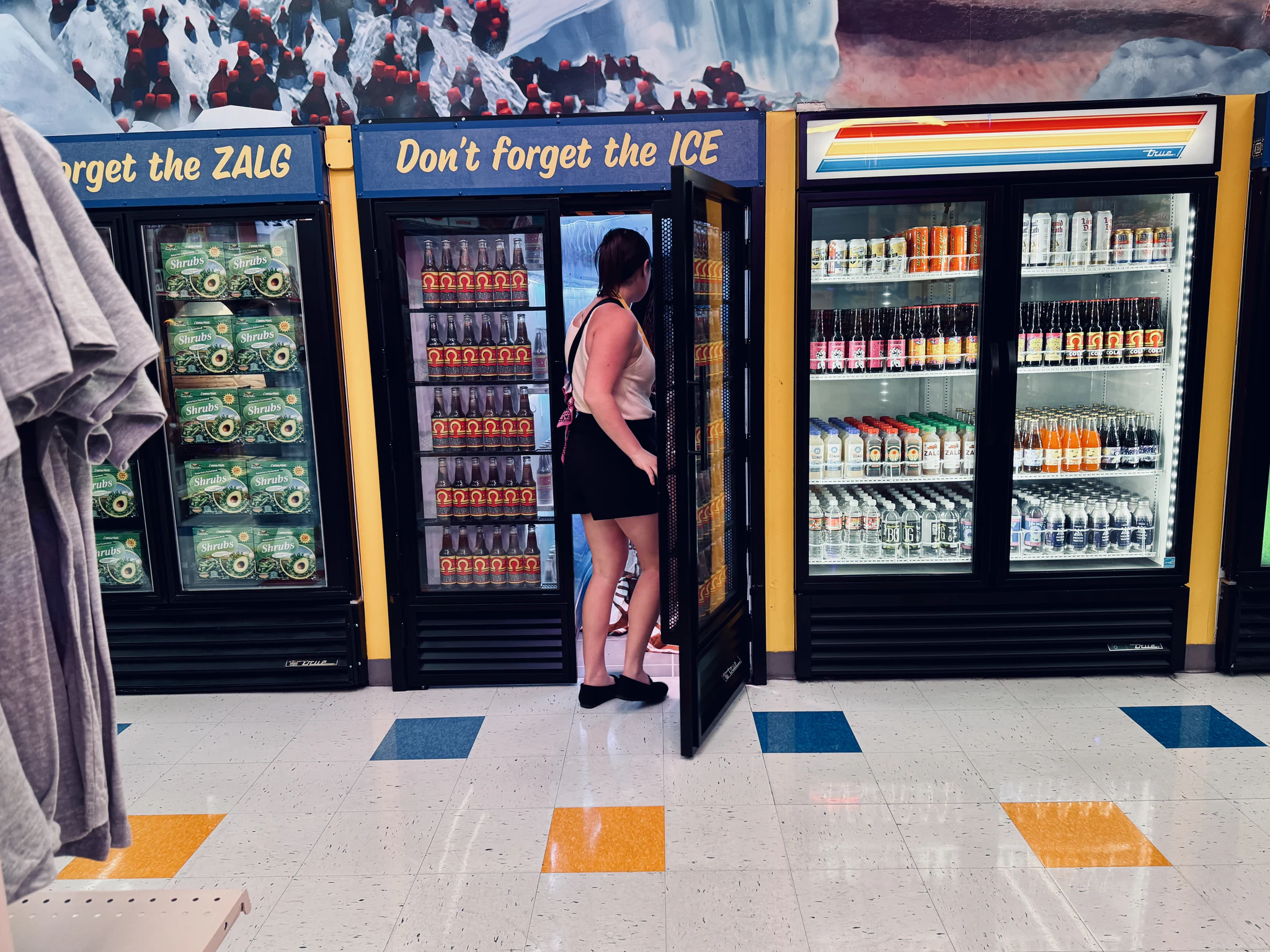Drinks fridges in Omega Mart signed Don't forget the ICE, a shopper reaching into one of them.