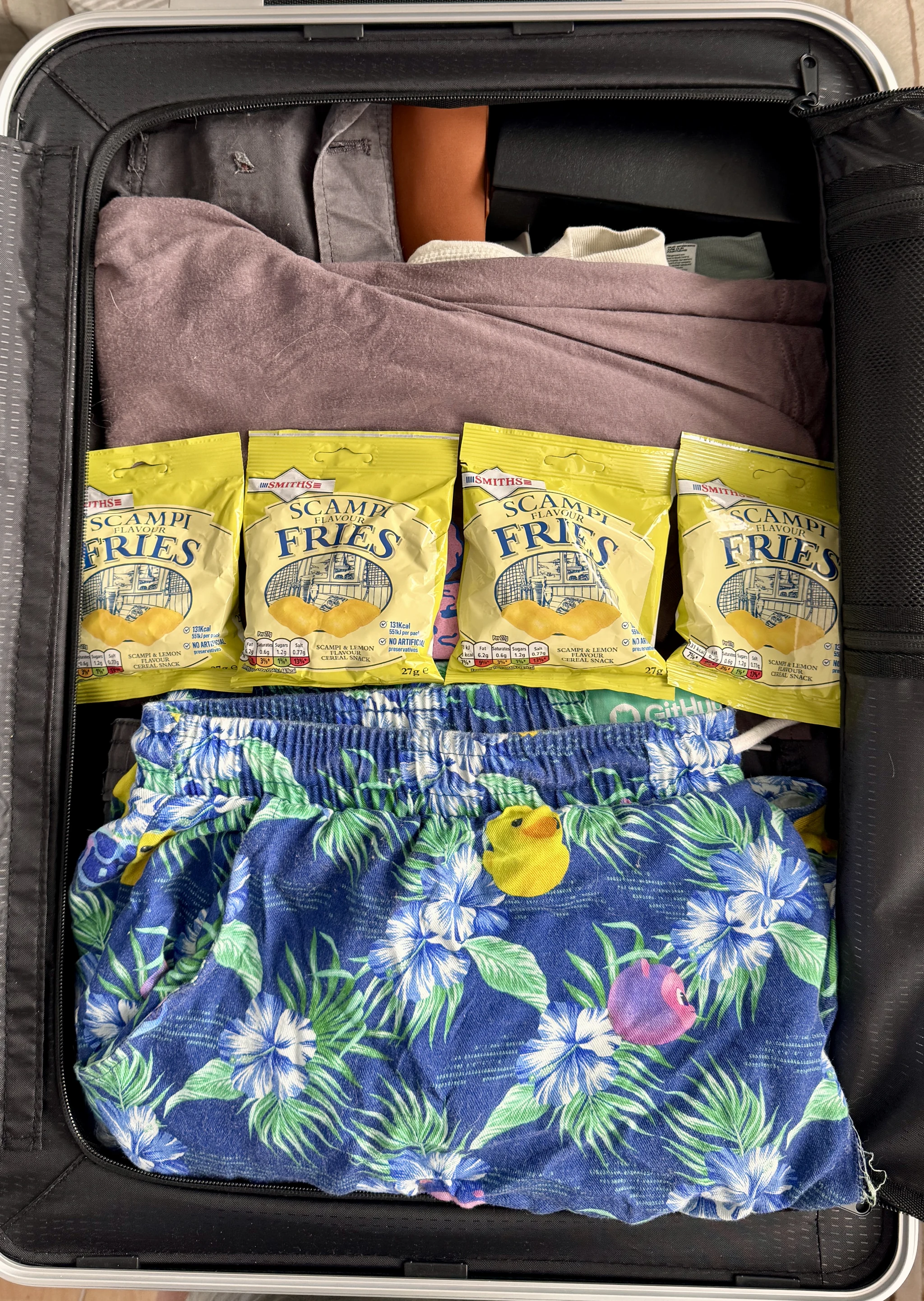 An open suitcase containing folded clothes, blue floral swimming shorts and four packets of Scampi Fries.