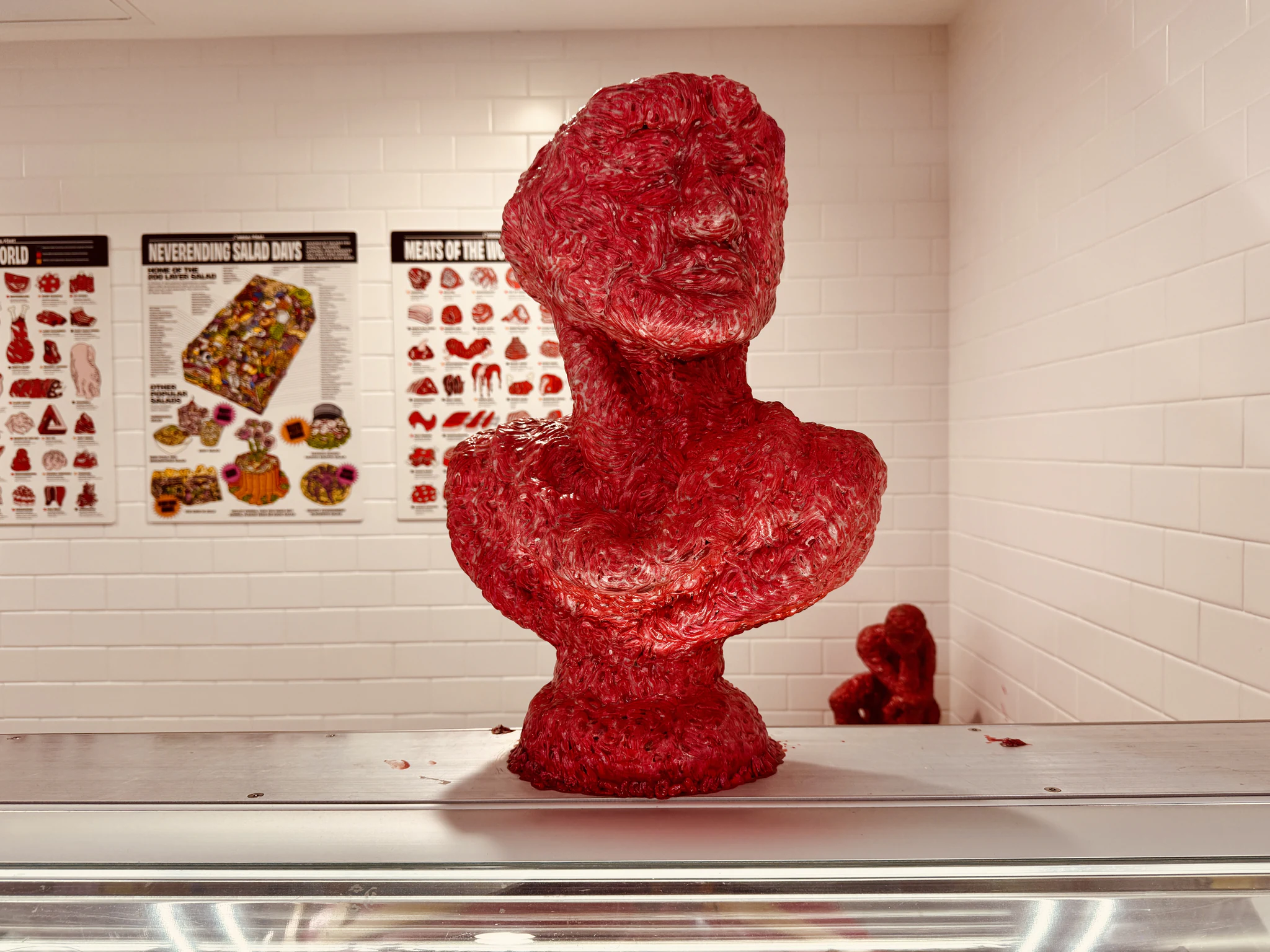 A bust sculpted from red processed meat on a deli counter, in front of posters titled Meats of the World.